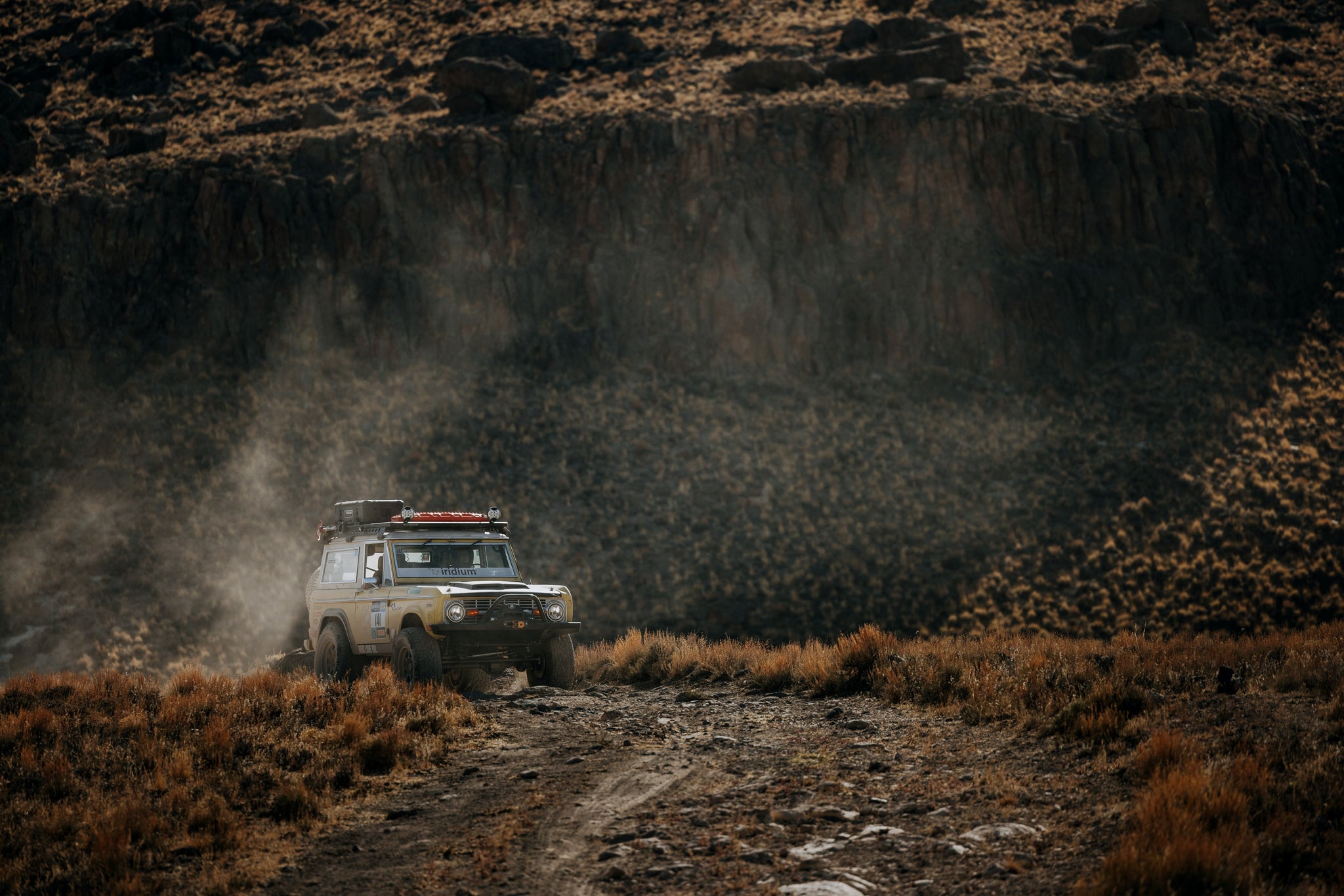 An off-road vehicle drives on a dusty dirt track through a rugged, dry mountain landscape with a large rocky cliff in the background.